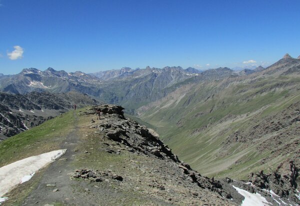 pianoro in alta quota con lembo di neve, rocce e radi prati, che si innalza su vallone sottostante. Due persone in secondo piano. intorno la catena montuosa