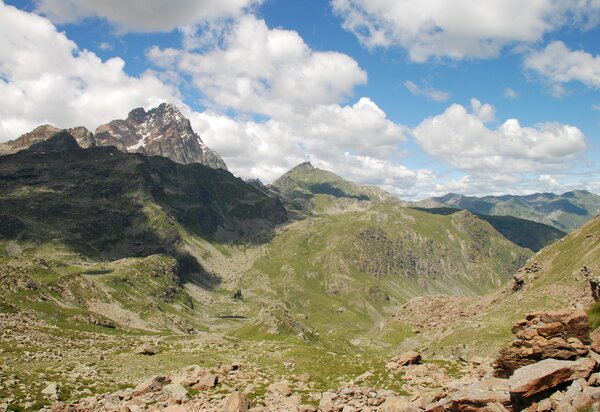 La cima del Monviso tra le nuvole sbuca dietro altre montagne coperte di pascoli. In primo piano alcune rocce