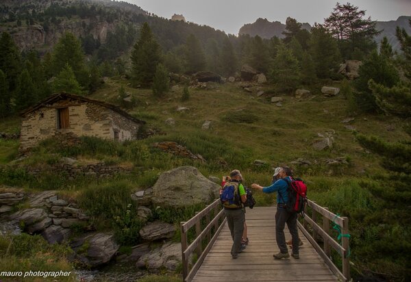 Escursionisti su un ponte di legno che supera il torrente vallanta; sulla sinistra una baita in pietra, sullo sfondo prato verde e larici
