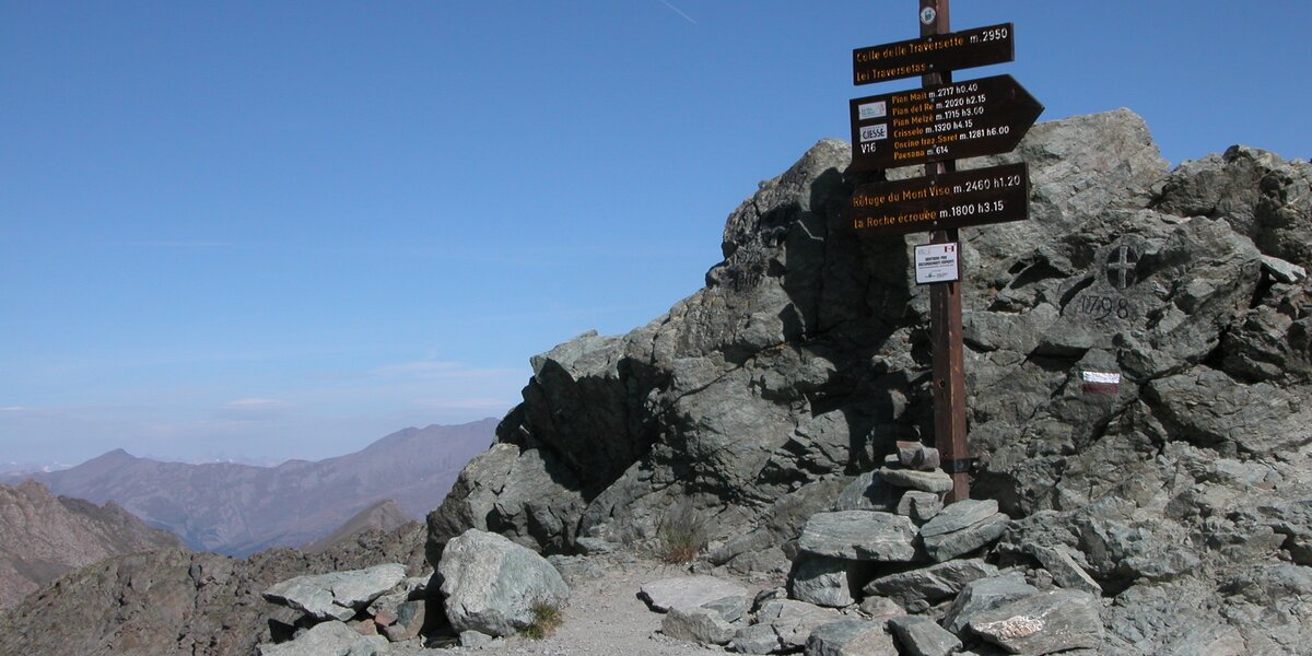 palina verticale di indicazione sentieristica addossata ad una roccia, sulla parte destra della foto; a sinistra lo sguardo scende verso la bassa valle