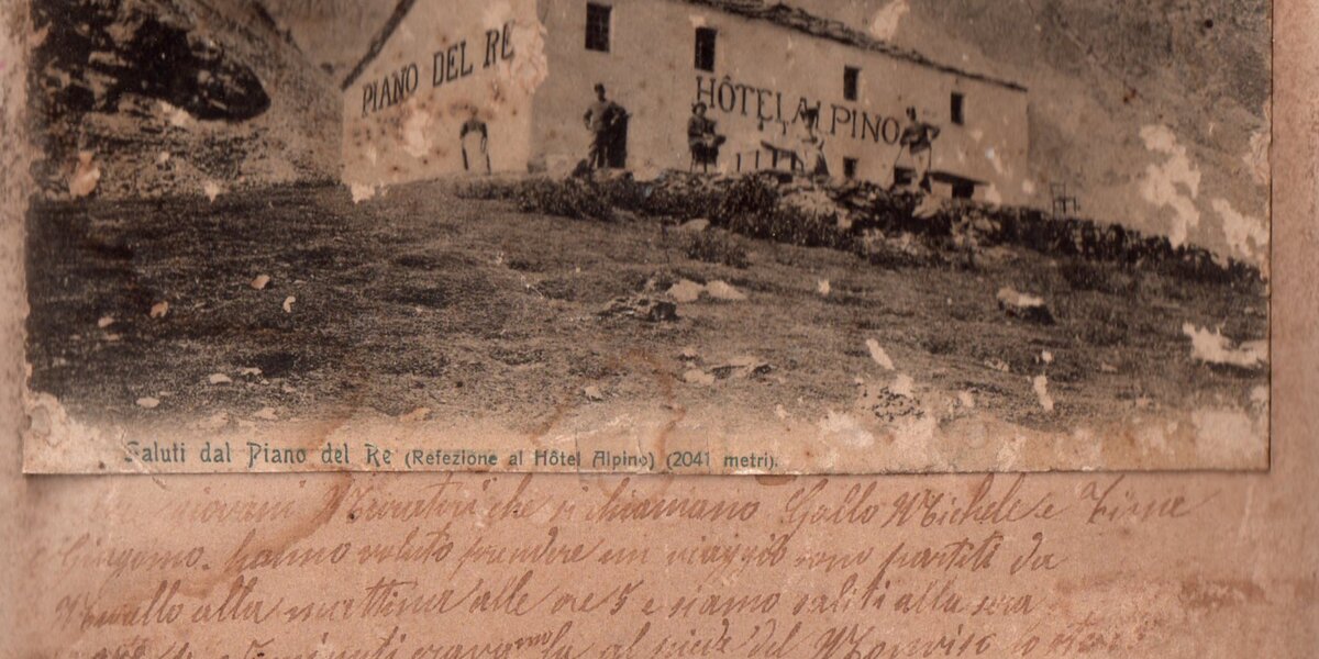 una vecchia fotografia del 1909 che raffigura l'albergo rifugio Pian del Re al tempo: un caseggiato bianco con l'insegna Pian del Re hotel alpino. Sotto alla foto, alcune righe di testo scritte a mano all'epoca.