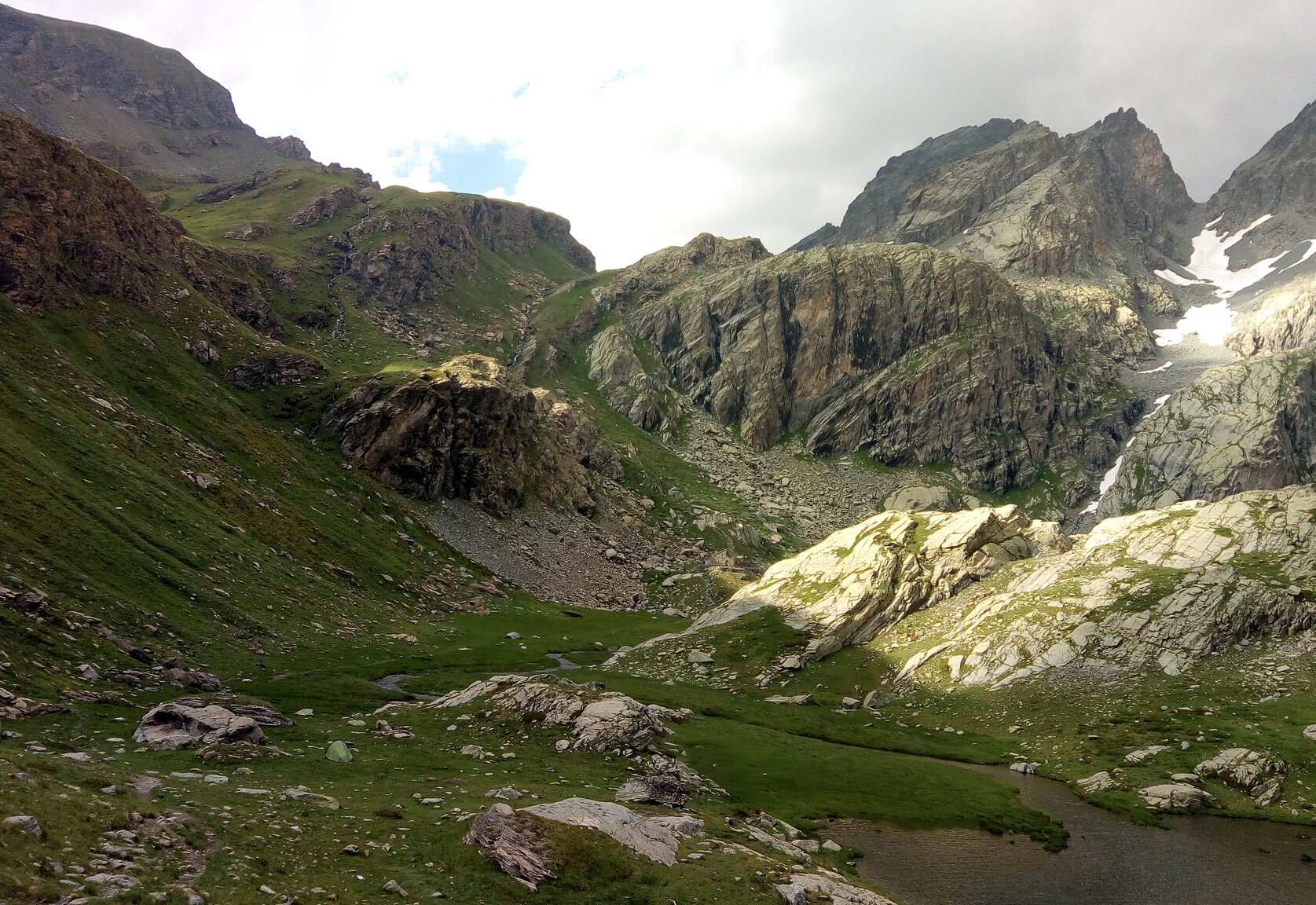 ambiente montano di alta quota con a sinistra prateria alpina e versanti inerbiti, a destra rocce e torrente che sfocia in un lago