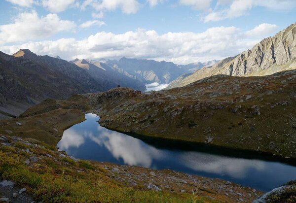 Un lago alpino dalla forma allungata in cui si specchiano le nuvole, intorno imponenti montagne, cielo azzurro.