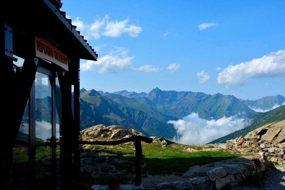A destra, in ombra, l'ingresso del rifugio Granero con vetrata e targa in legno. Dal rifugio piccola staccionata in legno lungo il sentiero pietroso. dietro panorama montano con la catena montuosa tra le nuvole.