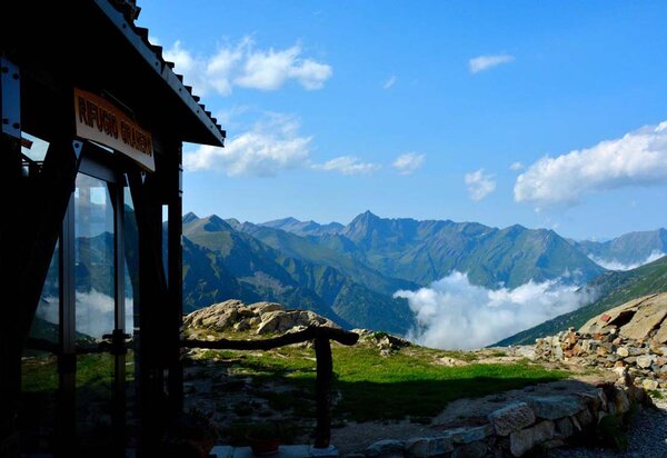A destra, in ombra, l'ingresso del rifugio Granero con vetrata e targa in legno. Dal rifugio piccola staccionata in legno lungo il sentiero pietroso. dietro panorama montano con la catena montuosa tra le nuvole.